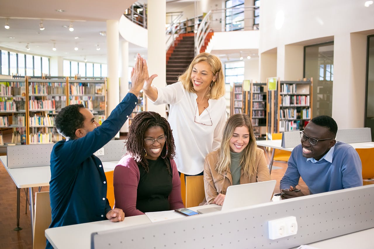 Positive multiracial classmates smiling happily and having high five while doing successful research on laptop together